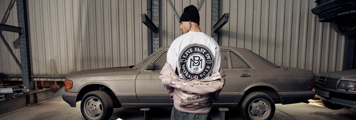 A person in a beanie stands with their back to the camera in a garage, wearing a white jacket with a large Live Fast logo, next to two dusty vintage cars.