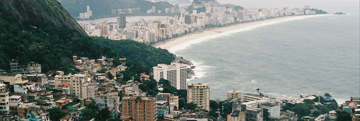 A panoramic view of Rio de Janeiro’s coastline, showing dense urban buildings, lush green hills, and a long sandy beach curving along the Atlantic Ocean under a cloudy sky.