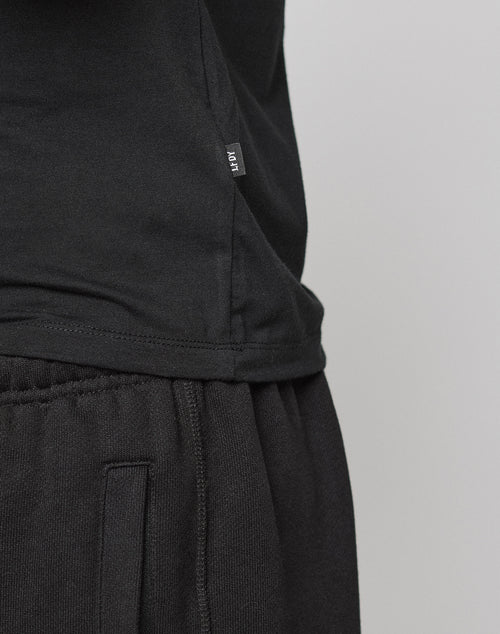 Close-up of a person in a black LFDY Slim Tee by LIVE FAST DIE YOUNG and black sweatpants. The side hem shows a small Flag-Label with white MOVT text. Made from soft Baumwolle, the tee is set against a plain light gray background.