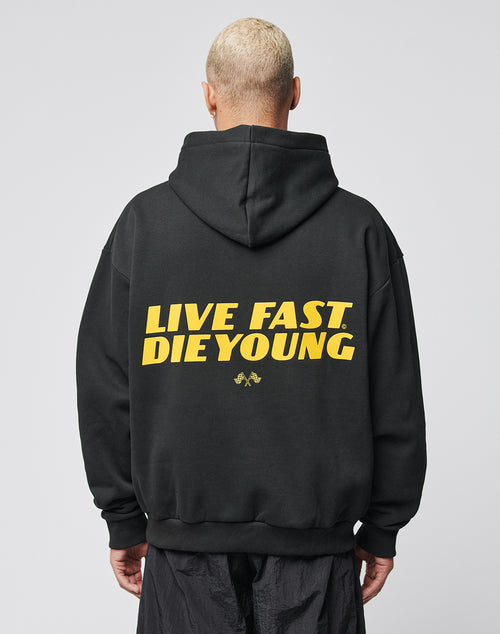 A person with short blond hair wears the LFDY Drift Hooded by LIVE FAST DIE YOUNG, a black hoodie with yellow LIVE FAST. DIE YOUNG text on the back, Oversize Fit, and doppellagige Kapuze, against a plain light background.
