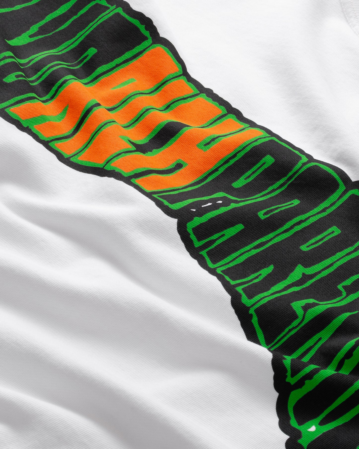A close-up of the LIVE FAST DIE YOUNG No Rich Parents Longsleeve, a white 100% Baumwolle regular fit shirt with bold green, black, and orange diagonal text; exact wording partly obscured.