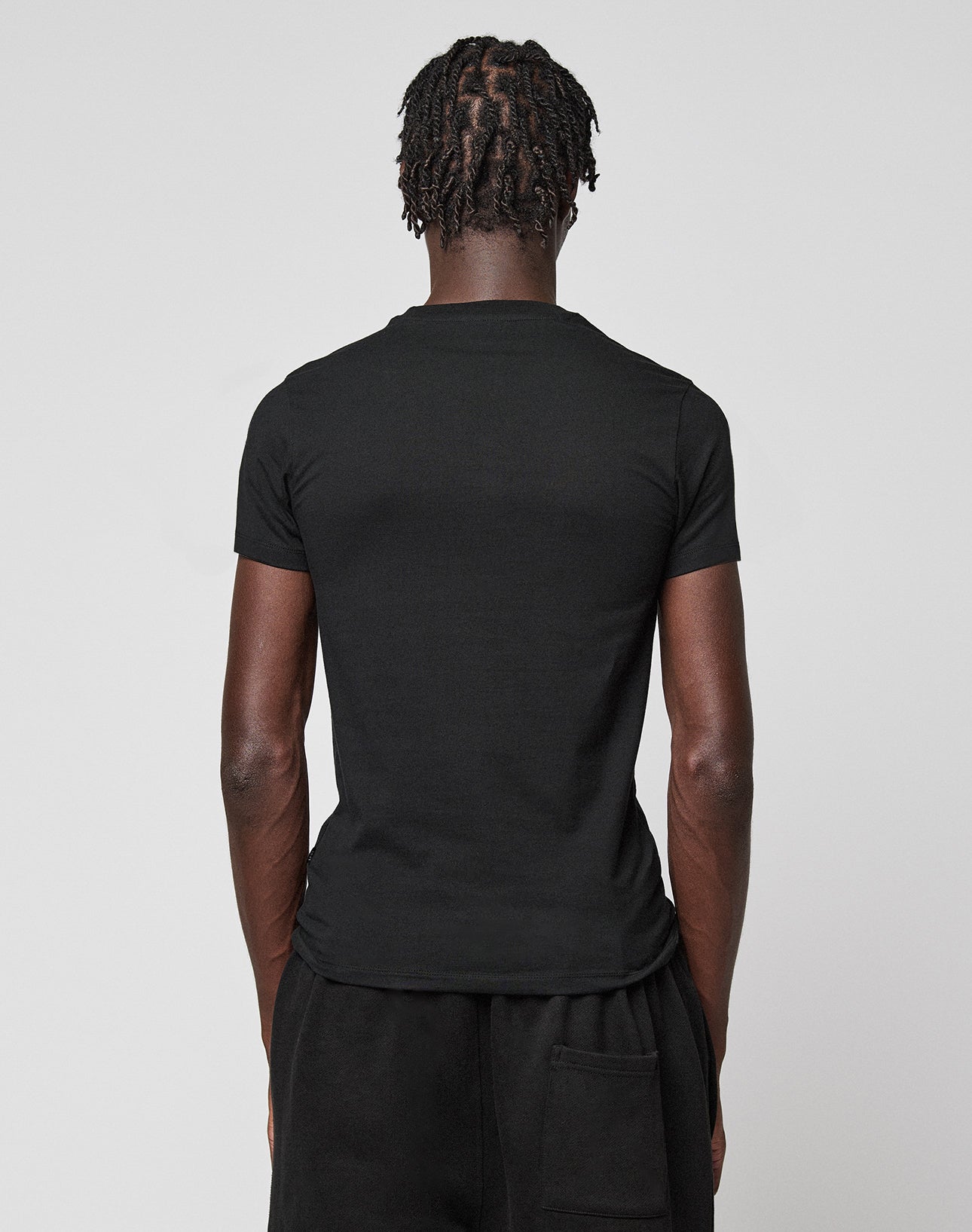 A person with short, twisted hair is shown from behind in a plain black LIVE FAST DIE YOUNG LFDY Slim Tee and black pants against a light gray background. A small Flag-Label detail completes the minimalist look.