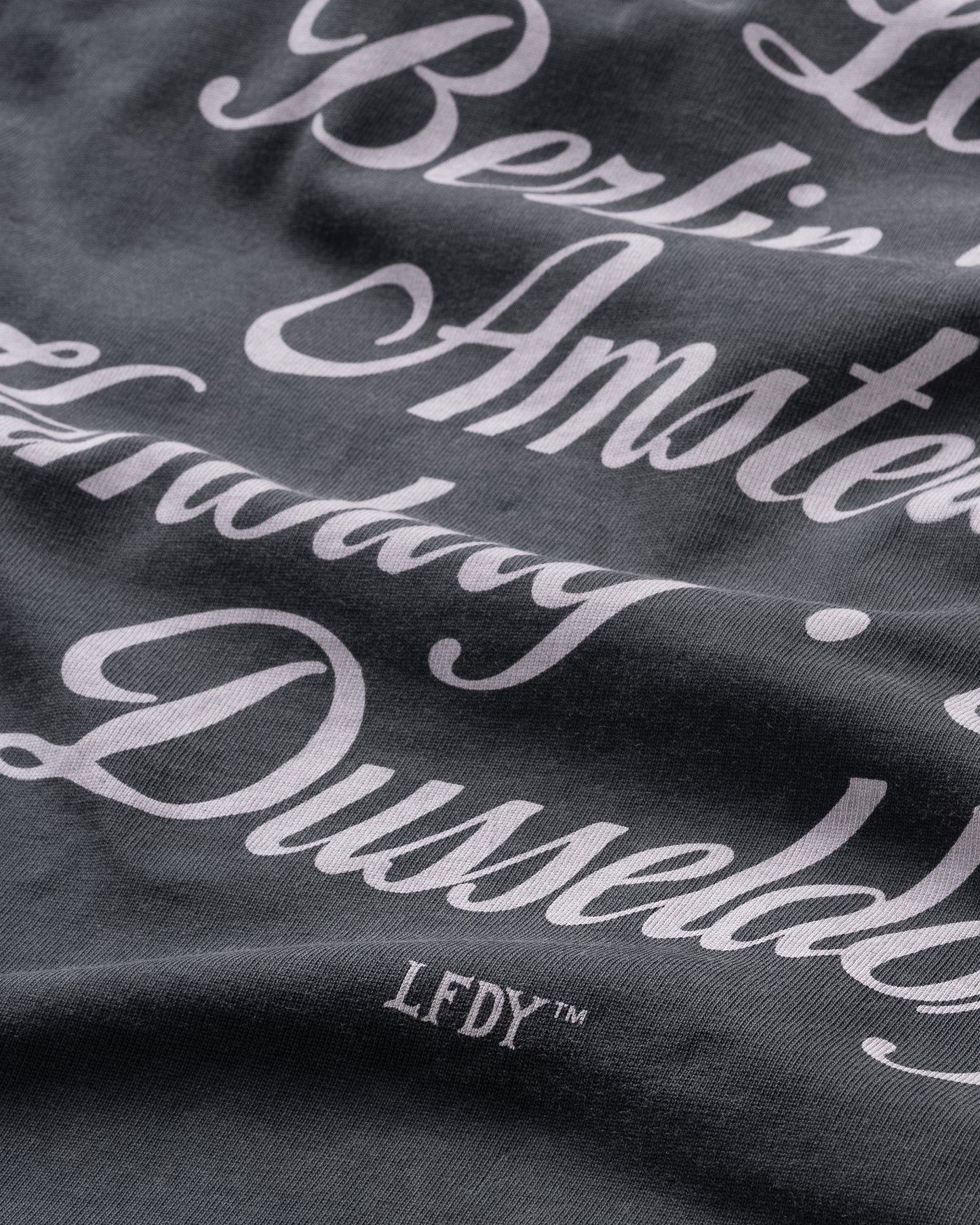 A close-up of the LIVE FAST DIE YOUNG Black Chrome Tee, featuring dark gray fabric with white cursive city names. The LFDY™ logo appears at the bottom. Crafted from 100% Baumwolle in a stylish oversize fit.