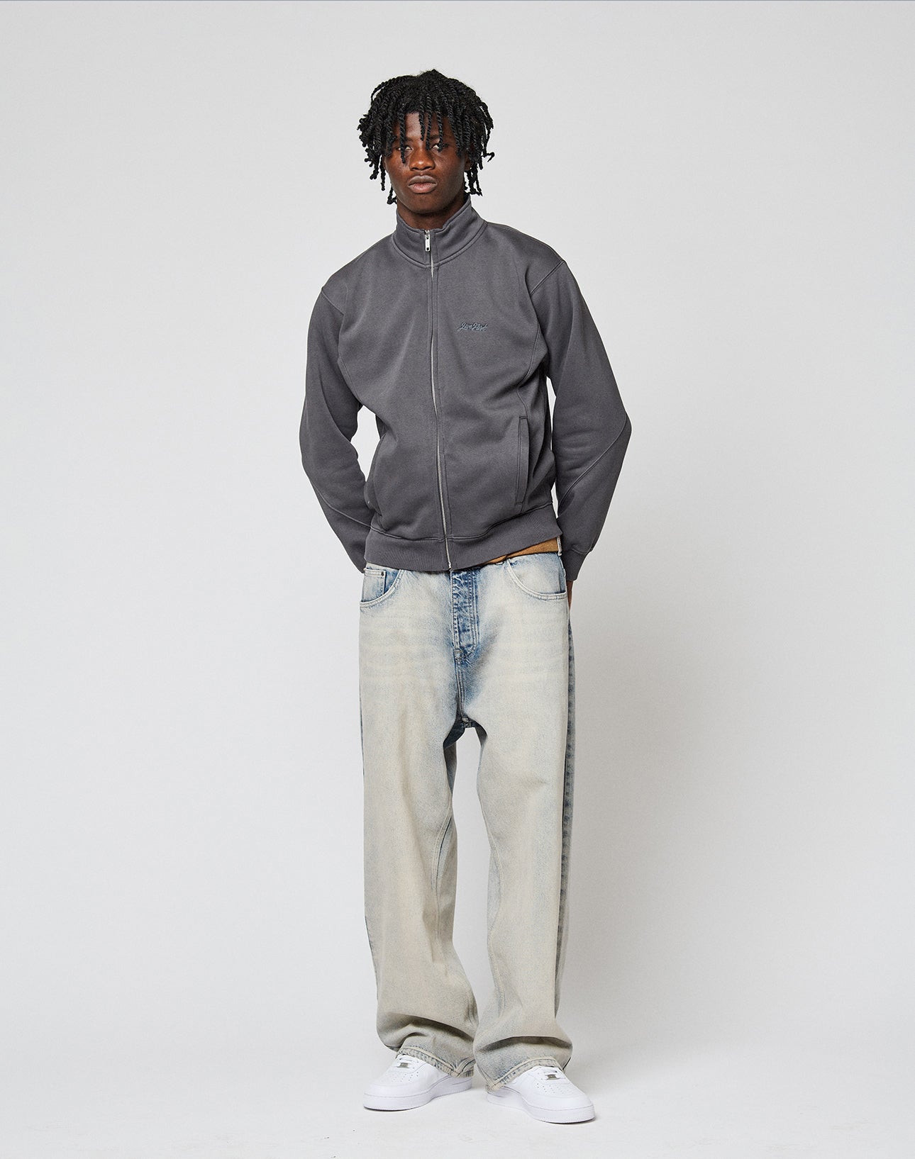A man with short dreadlocks wears a LIVE FAST DIE YOUNG Sweatsuit Jacket, light-wash regular fit baggy jeans, and white sneakers, standing against a plain light gray background with his hands in his pockets.