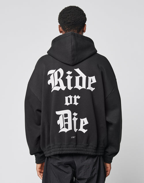 Someone stands against a plain background wearing the LIVE FAST DIE YOUNG Ride or Die Hooded Zip, a black oversize hoodie with doppellagige Kapuze and Ride or Die in bold white Gothic letters across the back.
