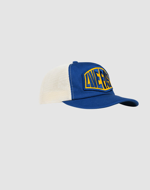 The LFDY Patched Trucker by LIVE FAST DIE YOUNG is a blue and white trucker cap with snap closure, curved brim, and a yellow-blue LIFERS logo patch on the front, shown against a plain light gray background.