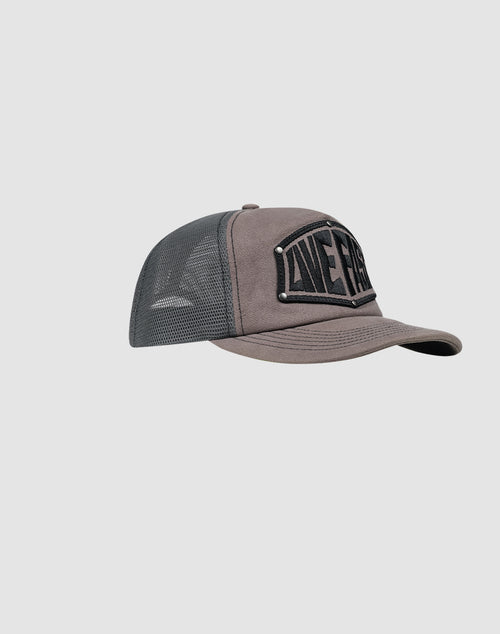 The LFDY Rivet Trucker cap by LIVE FAST DIE YOUNG features gray and black mesh, a curved brim, a bold rectangular front patch, and a subtle Flag-Label detail. The hat is shown at a slight left angle against a light background.