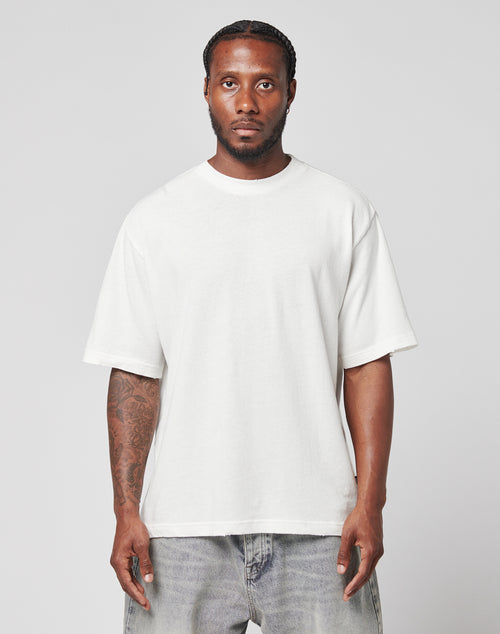 A man with braided hair and tattoos on his right arm wears the LIVE FAST DIE YOUNG LFDY Vintage Tee in an oversize fit with light blue jeans, standing against a plain light gray background, looking directly at the camera neutrally.