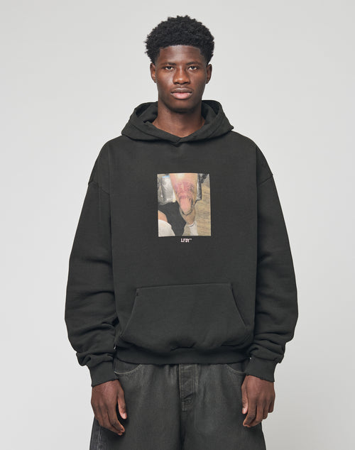 A man with short curly hair wears the LIVE FAST DIE YOUNG LFDY Sharkhunter Hooded in black, featuring a front graphic, standing against a plain light gray background.