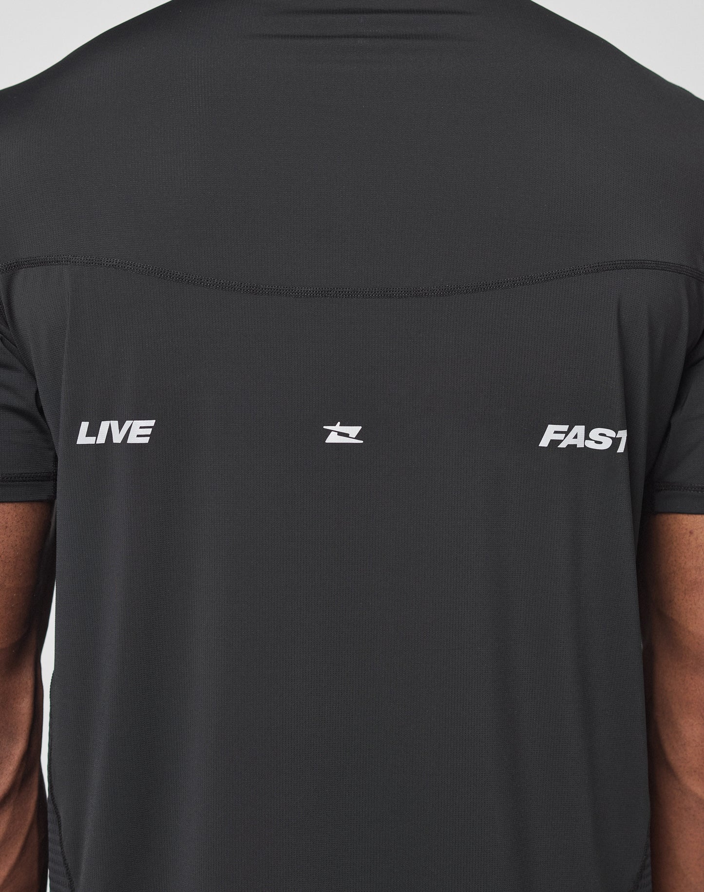 Back view of a person wearing the LIVE FAST DIE YOUNG Live Fast Performance Tee—a slim fit black Nylon Spandex shirt with LIVE on the left, FAST on the right, central logo, and reflective front/back prints.