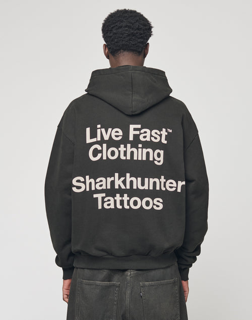 A person stands facing away, wearing the LFDY Sharkhunter Hooded by LIVE FAST DIE YOUNG. This black Oversize Fit hoodie is crafted from soft Baumwolle and features bold white text: Live Fast Clothing Sharkhunter Tattoos on the back.