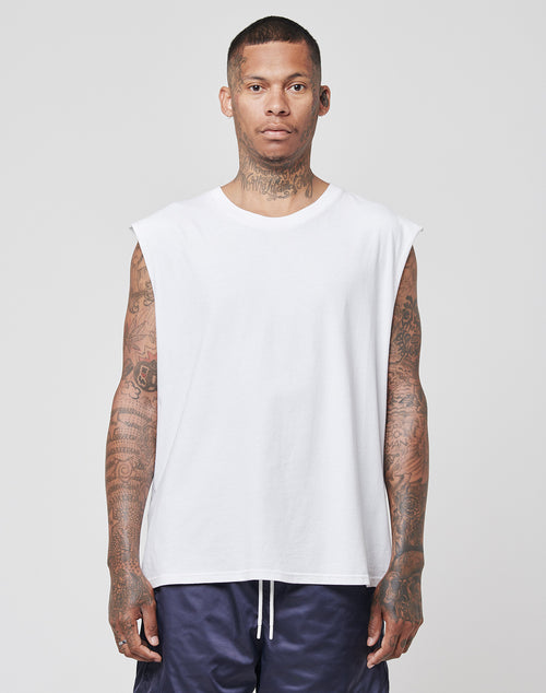 A tattooed man wears the LIVE FAST DIE YOUNG Basic Muscle Shirt, a loose-fit sleeveless white top made from 100% cotton, paired with dark shorts against a plain light gray background, looking straight at the camera.