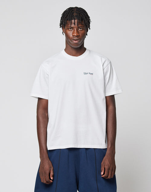 A man with short braided hair wears the LIVE FAST DIE YOUNG Live Fast Paris Tee, a regular fit 100% cotton white t-shirt with front and back print, and dark blue pants, standing against a gray background with a neutral expression.