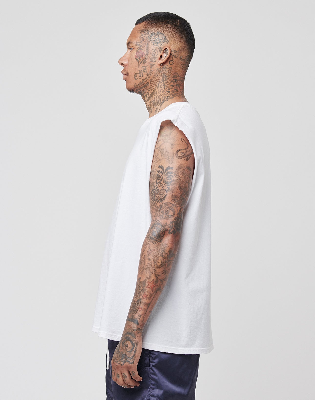 A tattooed man stands in profile against a plain white background, wearing the LIVE FAST DIE YOUNG Basic Muscle Shirt (100% Baumwolle, sleeveless, Loose Fit) in white and dark shorts.