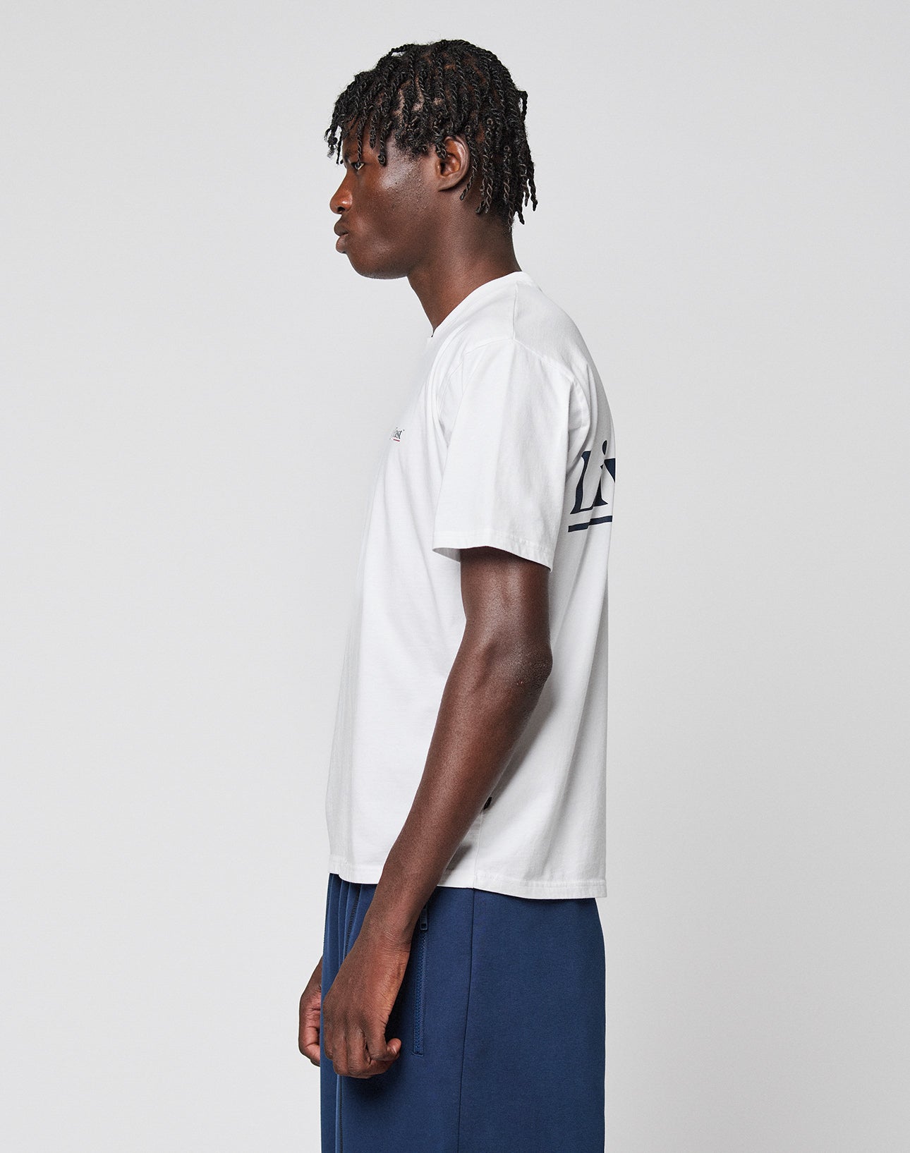 A person with short, textured hair stands in profile wearing the LIVE FAST DIE YOUNG Live Fast Paris Tee, a white regular fit t-shirt with navy blue front and back print, and dark blue pants, set against a plain light gray background.