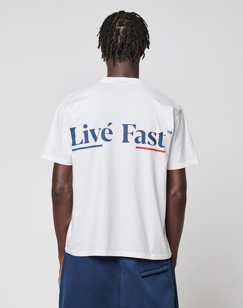 A person with short, twisted hair faces away, wearing the LIVE FAST DIE YOUNG Live Fast Paris Tee in white with bold Livé Fast™ blue front and back prints plus a red underline, styled with navy shorts. The tee is Regular Fit and made of 100% cotton.