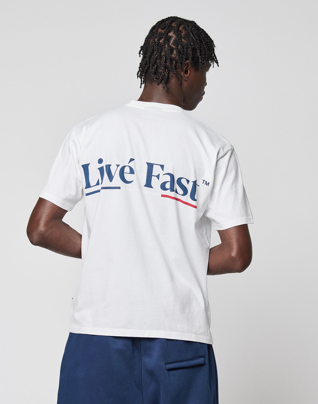 A person with short, twisted hair faces away, wearing the LIVE FAST DIE YOUNG Live Fast Paris Tee—100% Baumwolle, white, “Live Fast” in bold blue on the back with subtle front and back prints—paired with blue shorts against a plain light background.