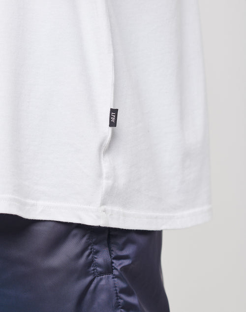 Close-up of a person wearing the LIVE FAST DIE YOUNG Basic Muscle Shirt, a loose fit white tee made from 100% Baumwolle with a small black flag-label on the hem, paired with dark shiny blue pants. Plain light background.