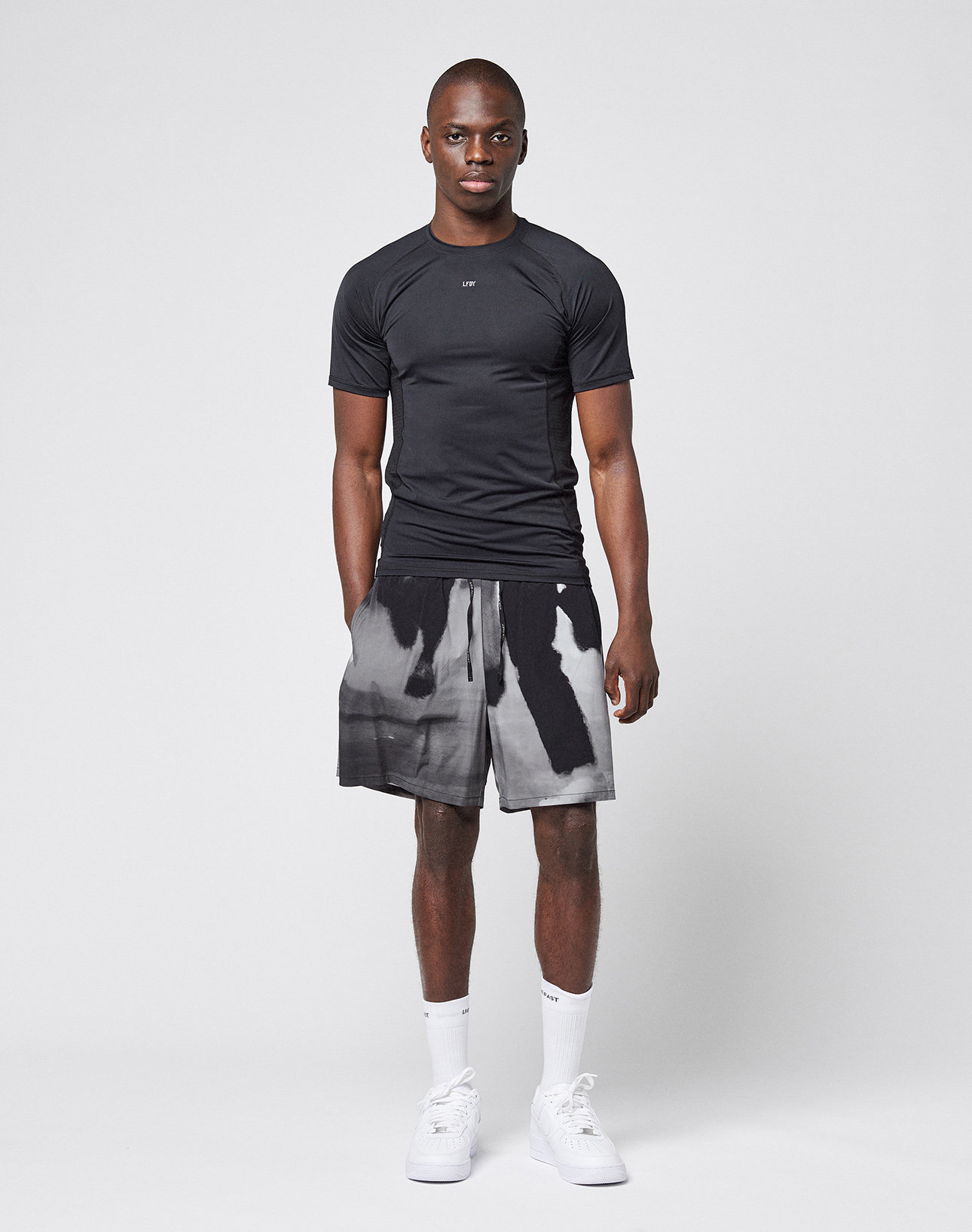 A man faces forward against a plain background, wearing the LFDY Performance Slim Tee by LIVE FAST DIE YOUNG with a reflective logo print, black and white tie-dye shorts, white crew socks, and white sneakers. His arms rest at his sides.