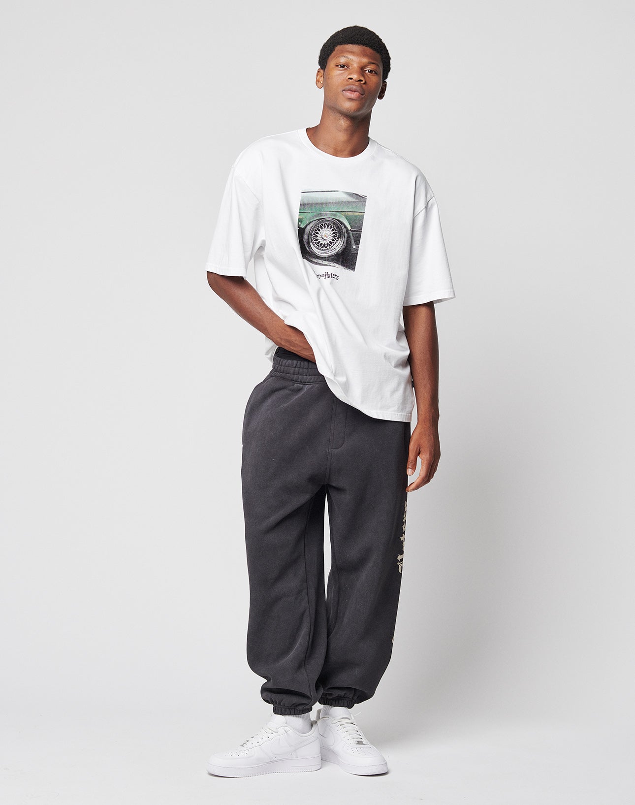A young man poses casually against a plain background, wearing a white graphic T-shirt, white sneakers, and the LIVE FAST DIE YOUNG LFDY Haters Jogger—wide fit dark sweatpants with an elastic waistband and pockets. One hand rests in his pocket as he looks upward.