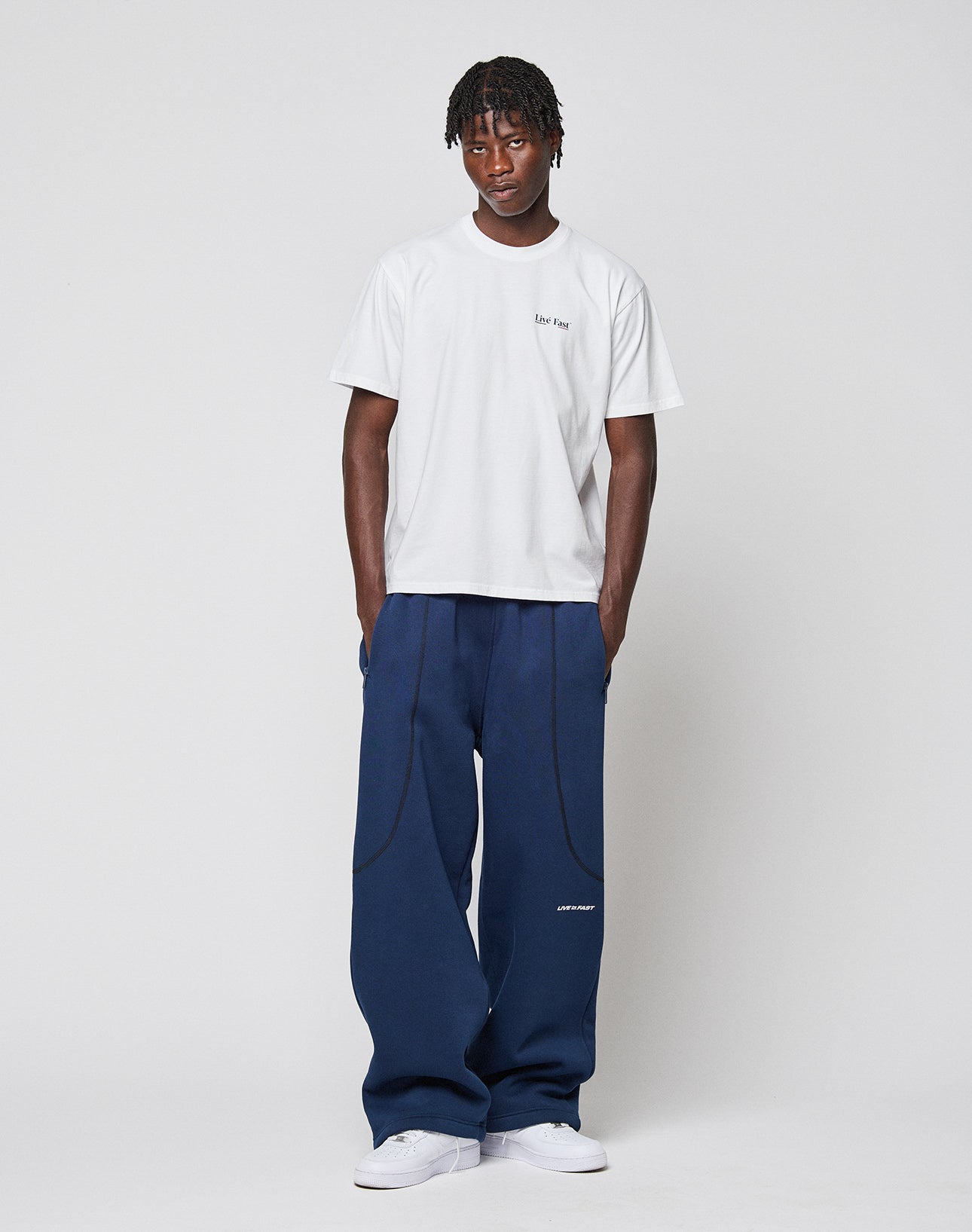 A person with short, twisted hair stands against a plain background, hands in pockets, wearing the LIVE FAST DIE YOUNG Live Fast Paris Tee (regular fit, 100% Baumwolle), loose navy pants, and white sneakers.