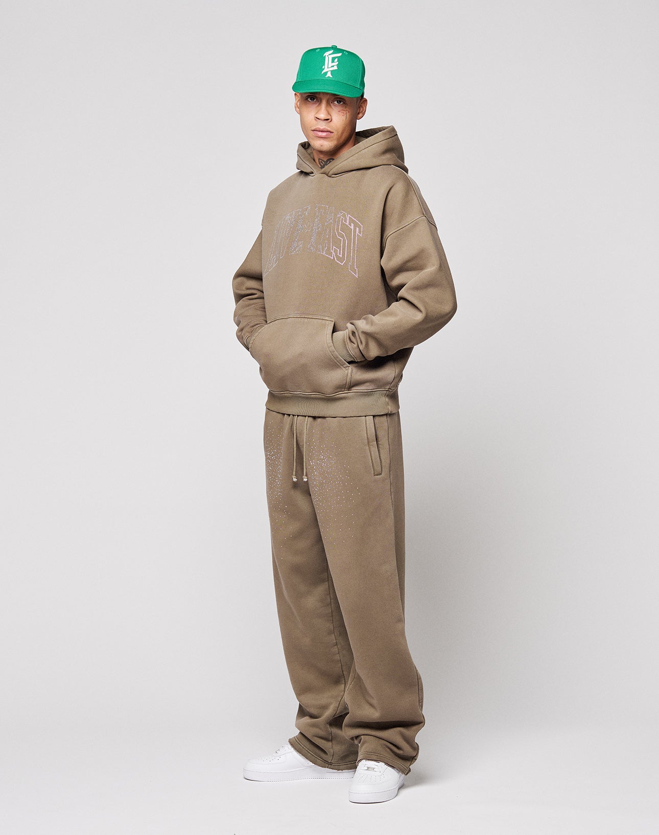 A person faces forward in the LIVE FAST DIE YOUNG Live Fast Rhinestone Hooded, styled oversized and brown, with matching sweatpants, a green baseball cap, and white sneakers, against a plain light gray background.