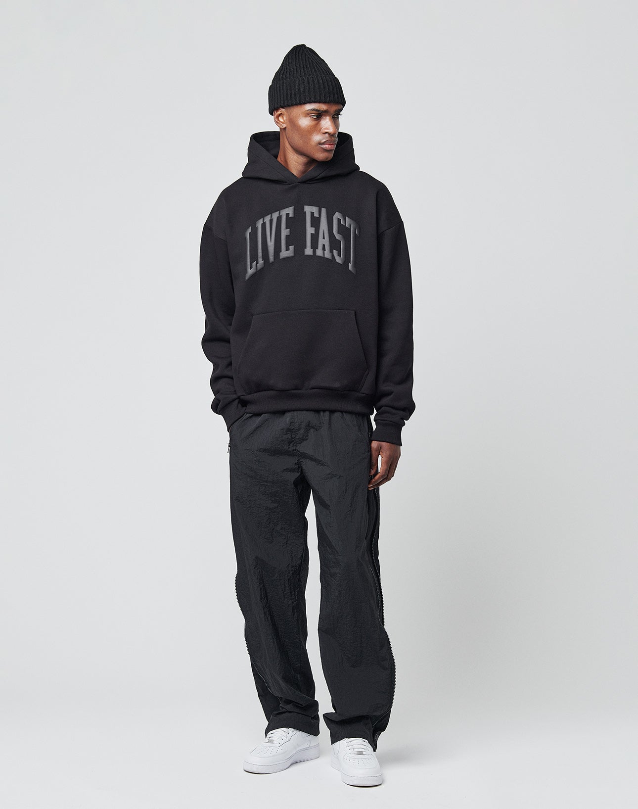 A man wearing a black LIVE FAST DIE YOUNG Live Fast College Hooded hoodie in an oversized fit, black beanie, black pants, and white sneakers stands against a plain light gray background.