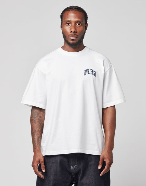 A man stands against a plain background, wearing the LIVE FAST DIE YOUNG Live Fast London Tee, an oversize fit 100% cotton white T-shirt with a front print, paired with dark baggy pants. He looks directly at the camera with a neutral expression.