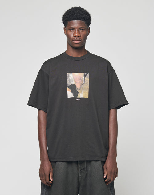 A man with short curly hair wears the oversize fit LFDY Sharkhunter Tee by LIVE FAST DIE YOUNG, featuring a rectangular graphic and LIFE on the front, paired with dark pants, standing against a plain light gray background.