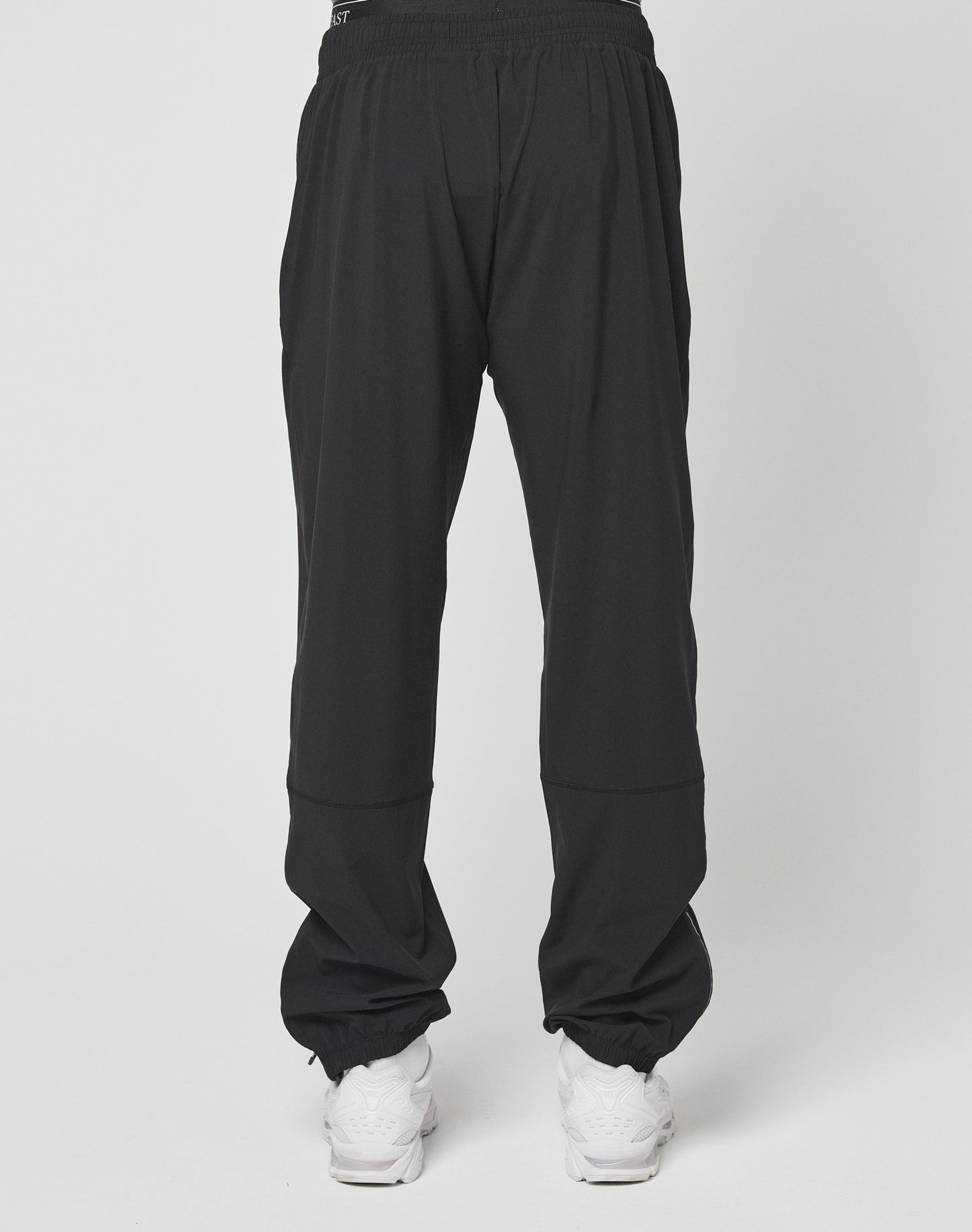 A person wearing LIVE FAST DIE YOUNG Live Fast Performance Running Pants in an oversize fit with an elastic waistband and white sneakers stands against a plain background, with the image focused on the back of the pants.