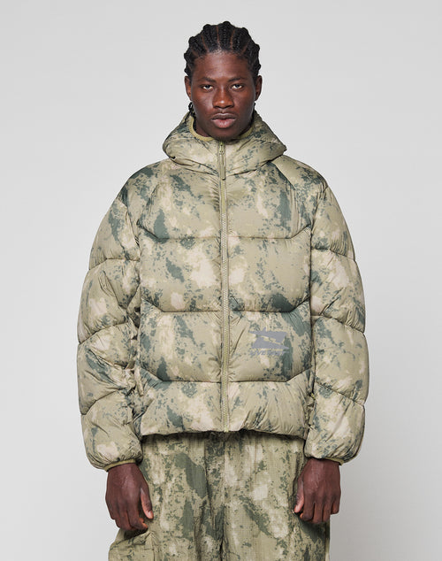 A person with braided hair wears the LIVE FAST DIE YOUNG Live Fast Puffer Jacket in green and beige camouflage with matching oversize-fit pants, standing against a plain light background.