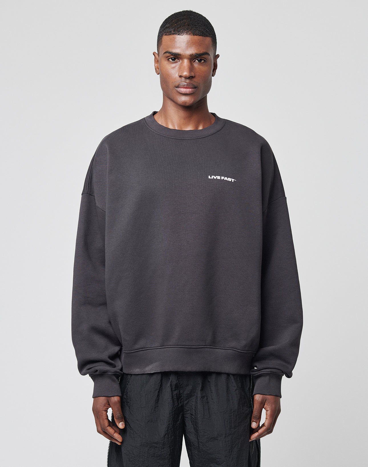 A man stands facing forward against a plain background, wearing a black LFDY Cinema Sweater by LIVE FAST DIE YOUNG with LIVE FAST. in small white letters on the upper left chest, paired with black pants.
