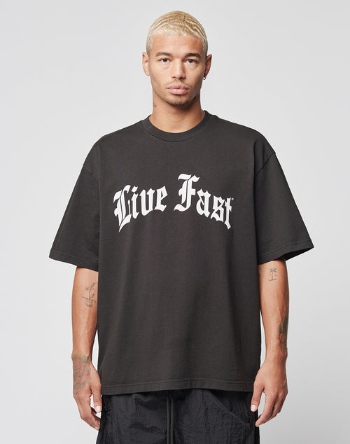 A person with short, bleached blonde hair wears the LIVE FAST DIE YOUNG Live Fast Fractured Tee—an oversized black cotton T-shirt with large white gothic “Live Fast” frontprint—standing against a plain white background.