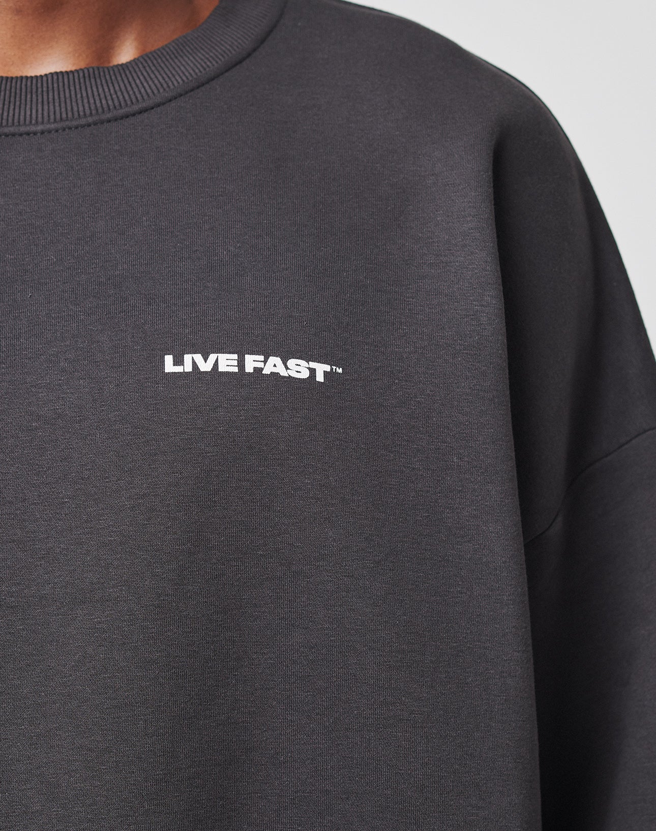 A close-up shows a person wearing the LFDY Cinema Sweater by LIVE FAST DIE YOUNG, featuring LIVE FAST™ in white on the chest. Only the shoulder and part of the neck are visible. Made from premium cotton-polyester.
