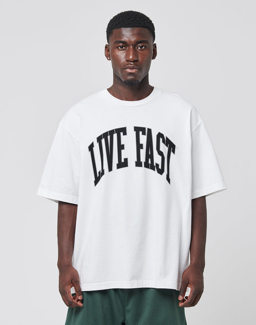 A man wearing the LIVE FAST DIE YOUNG Live Fast College Tee—a white, oversize cotton T-shirt with a bold LIVE FAST front print—stands against a plain gray background, looking directly at the camera.