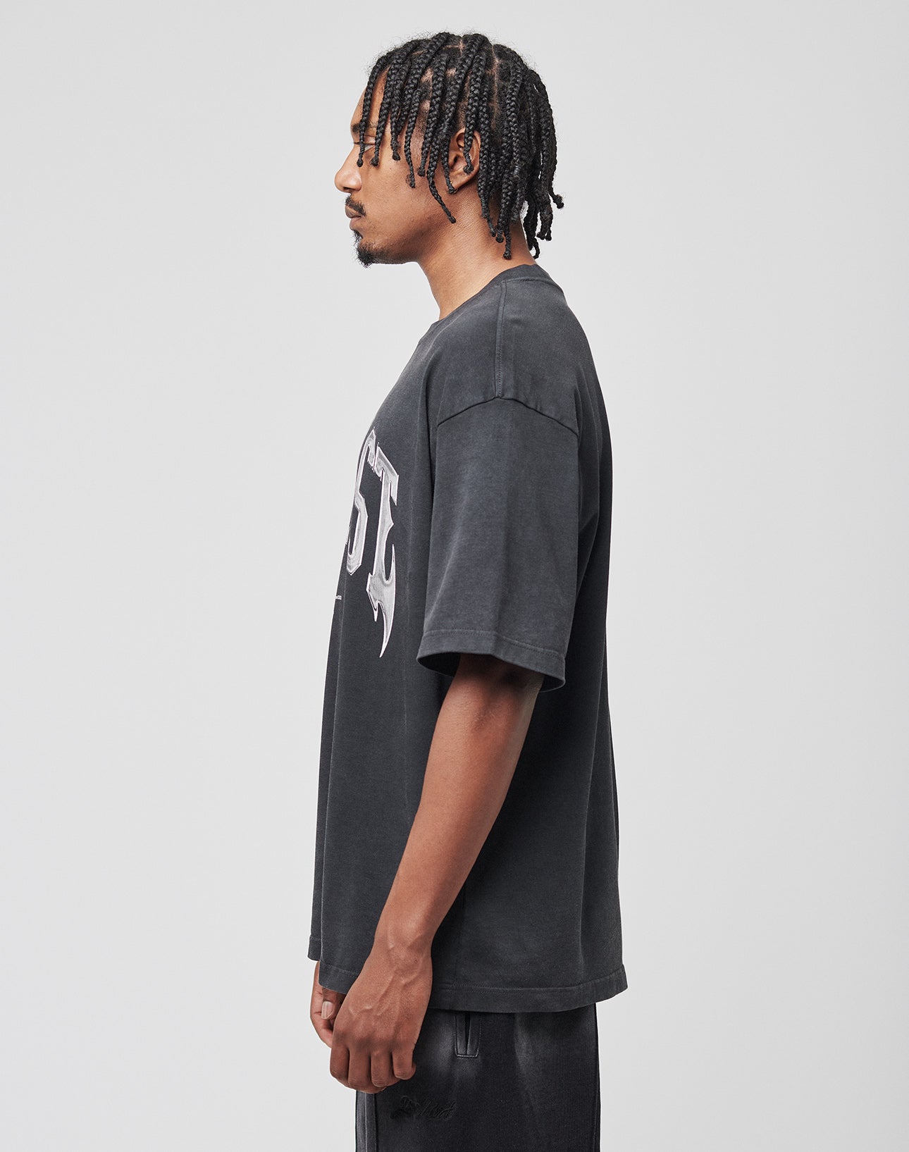 A man with braided hair faces left in profile, wearing the LIVE FAST DIE YOUNG Live Fast Axe Tee—an oversize fit, faded black T-shirt with a bold front print—and black shorts against a plain white background.