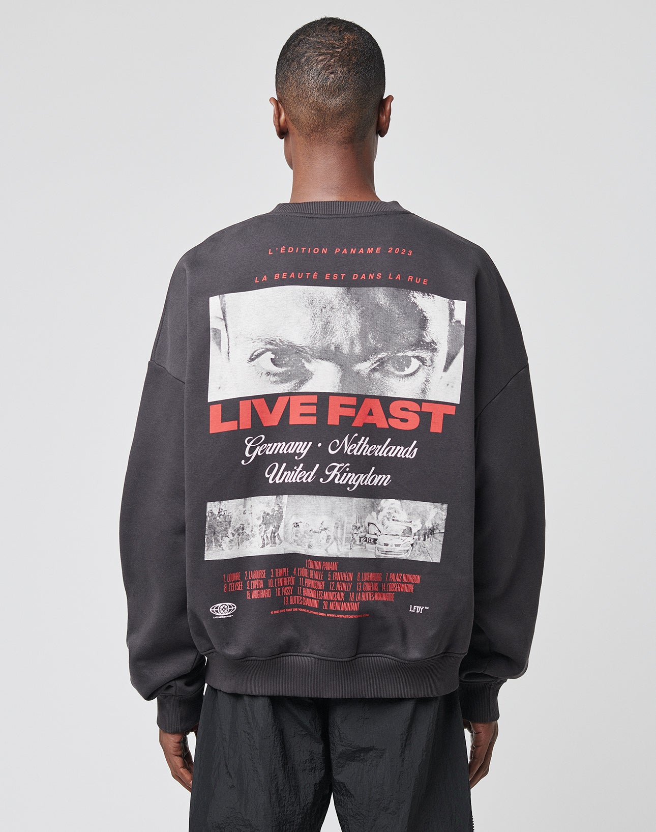 A person wears the LFDY Cinema Sweater by LIVE FAST DIE YOUNG, an oversize black Baumwolle-Polyester sweatshirt with bold LIVE FAST text, intense black and white eyes, and country names like Germany, Netherlands, and UK.