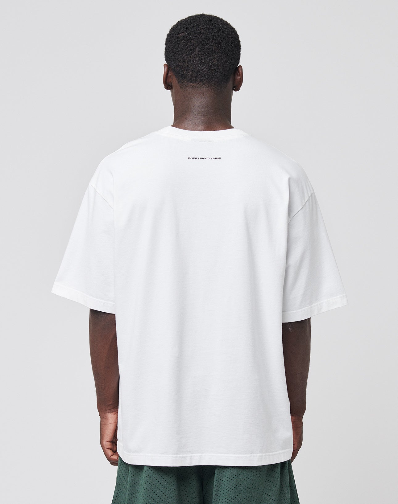 A person faces away from the camera wearing an oversize fit white Live Fast College Tee by LIVE FAST DIE YOUNG and dark green shorts, set against a plain light gray background.