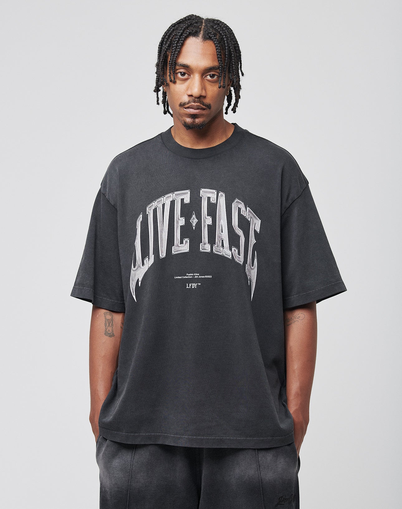 A man with short braids, mustache, and goatee faces the camera wearing the LIVE FAST DIE YOUNG Live Fast Axe Tee in black (Oversize Fit, Baumwolle) with a bold white LIVE FAST front print, paired with dark pants against a plain gray background.