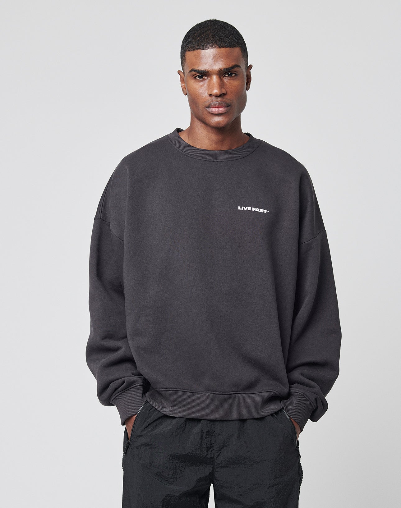 A man stands facing forward, wearing the LFDY Cinema Sweater by LIVE FAST DIE YOUNG—a black, oversized cotton-polyester sweatshirt with LIVE FAST in small white letters on the chest—and black pants. He looks neutral with his hands in his pockets.