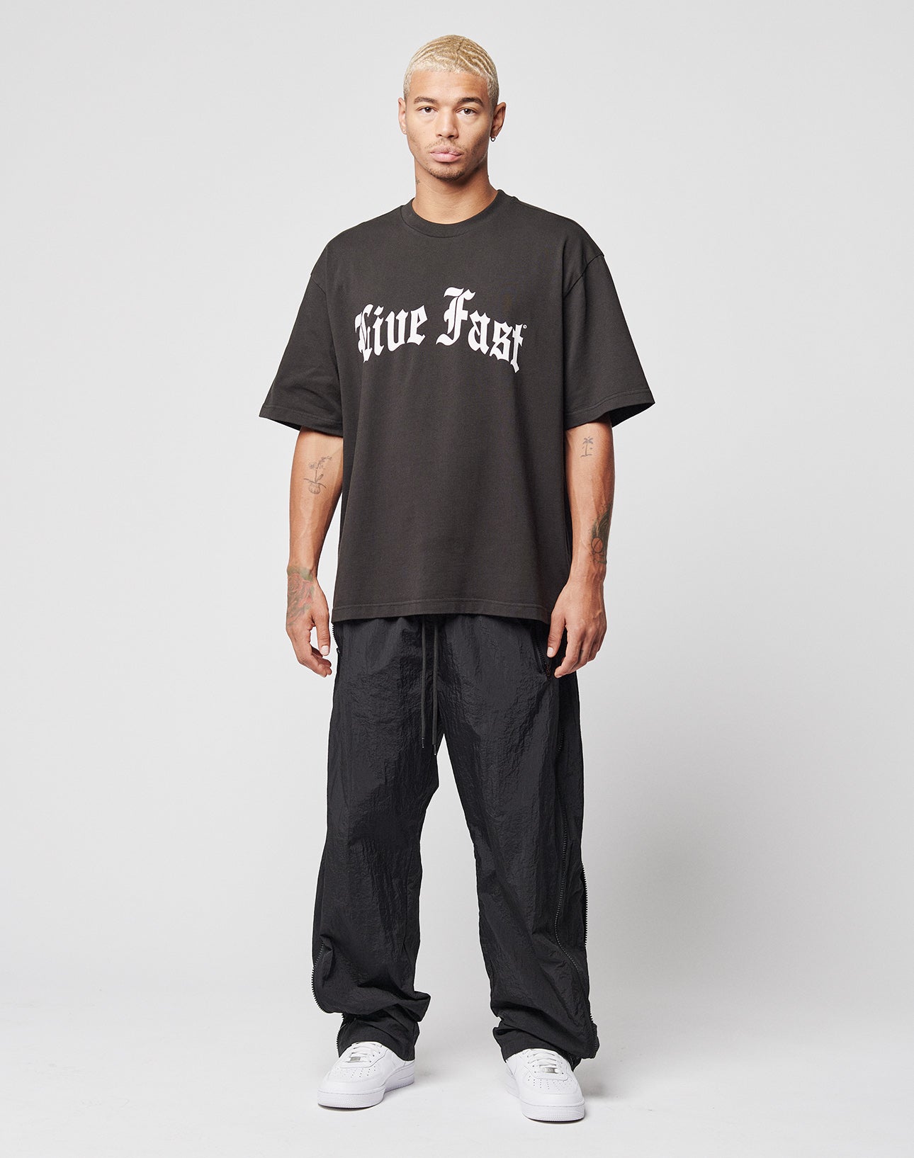A man with short blonde hair faces forward, wearing the LIVE FAST DIE YOUNG Live Fast Fractured Tee—an oversize black T-shirt with a bold Live Fast gothic front print—paired with black baggy pants and white sneakers against a plain white background.