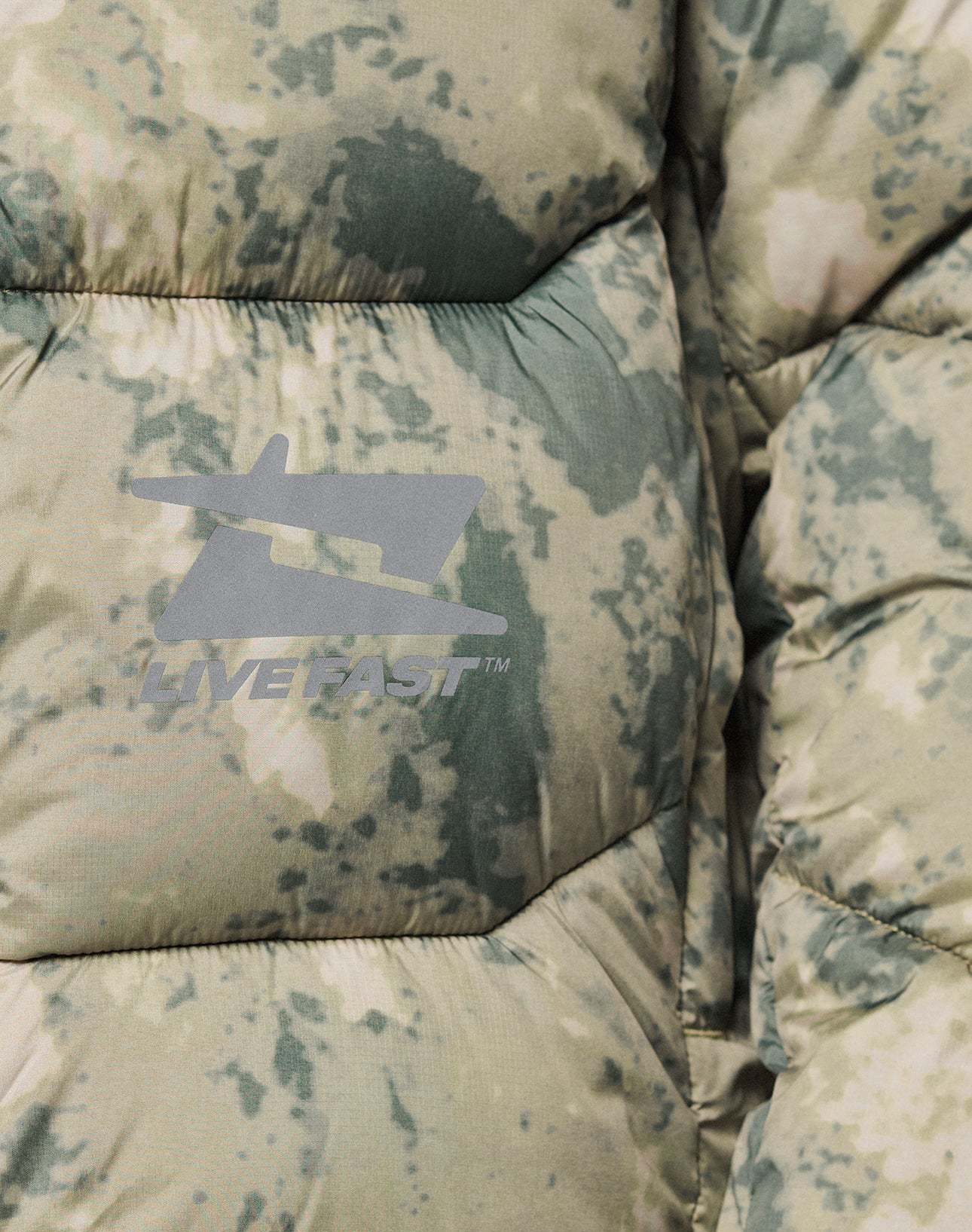 Close-up of the Live Fast Puffer Jacket by LIVE FAST DIE YOUNG, featuring a camo pattern, grey LIVE FAST™ logo, stylized airplane graphic on wasserabweisend fabric, oversize fit, and reflektierender Print for standout style.