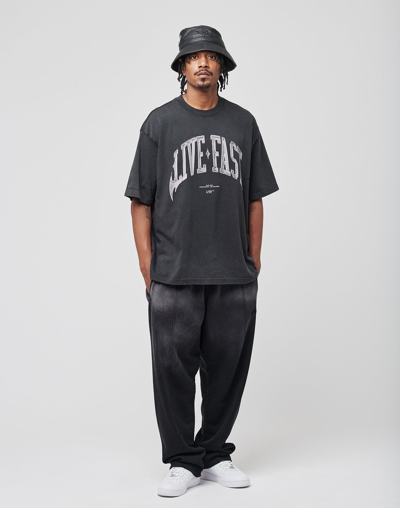 A man stands facing forward against a light gray background wearing the LIVE FAST DIE YOUNG Live Fast Axe Tee (an oversized black t-shirt with bold front print), baggy black pants, a black bucket hat, and white sneakers.