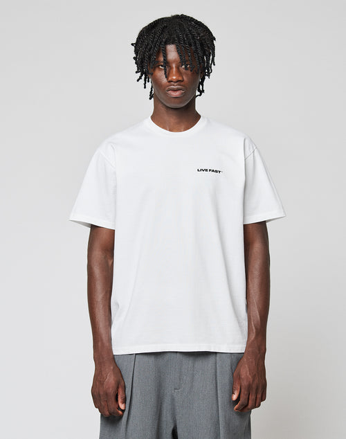 A person with short dreadlocks wears the LIVE FAST DIE YOUNG Live Fast Plain Tee, a regular fit white t-shirt with LIVE FAST. in small black letters on the left chest, made from 100% Baumwolle, paired with loose gray pants against a light background.