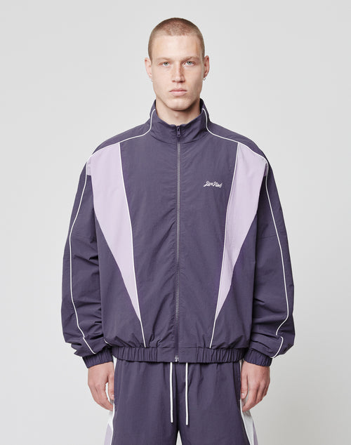 A person stands facing forward against a light gray background, wearing the LIVE FAST DIE YOUNG Basic Nylon Trackjacket in purple and lavender with matching pants. The regular fit features white piping, side seam pockets, and a Lost Art logo on the left chest.