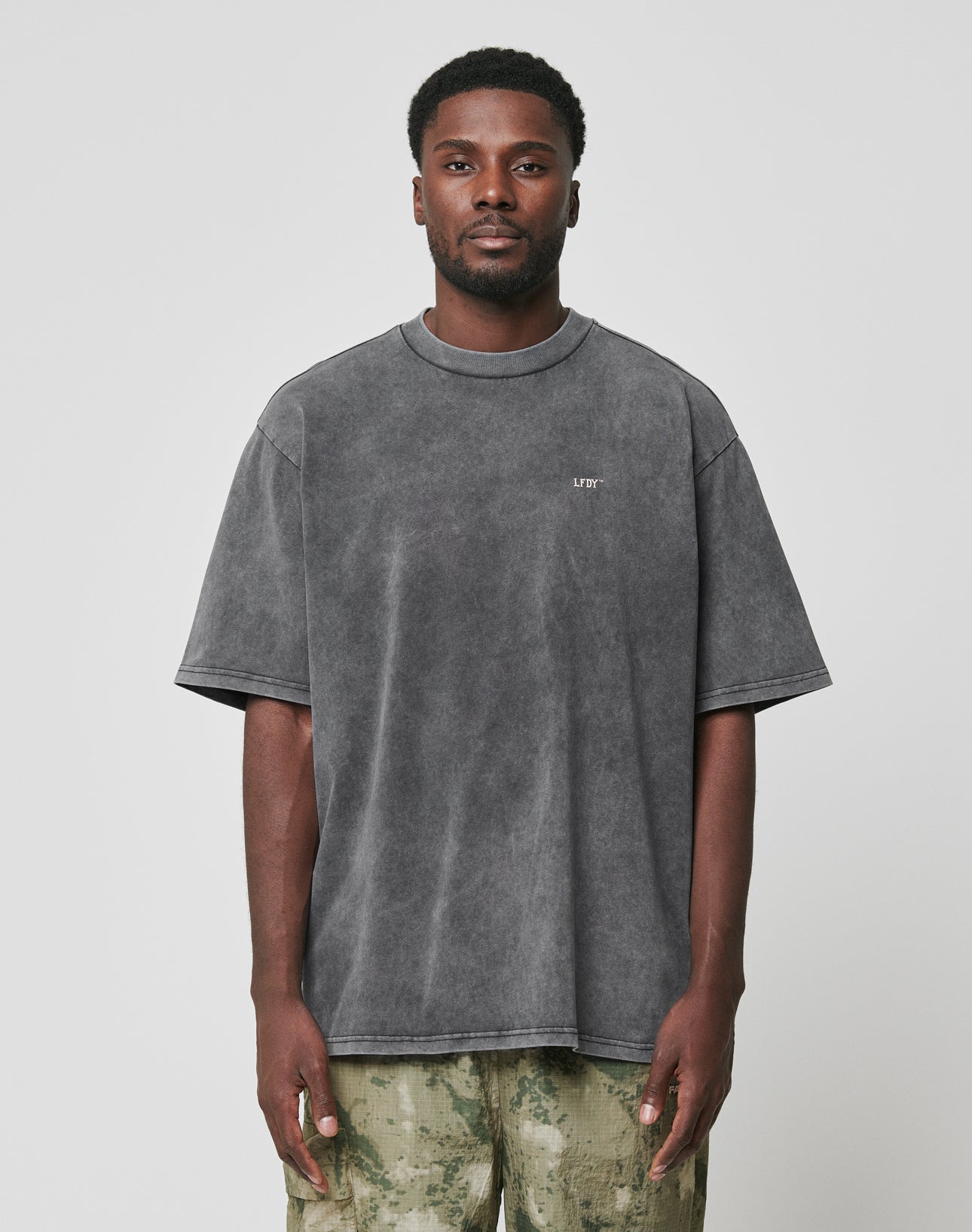 A man stands facing forward in an oversized, faded dark gray LIVE FAST DIE YOUNG LFDY Property Tee made from 100% Baumwolle, with short sleeves, paired with light green tie-dye pants. The background is plain white.