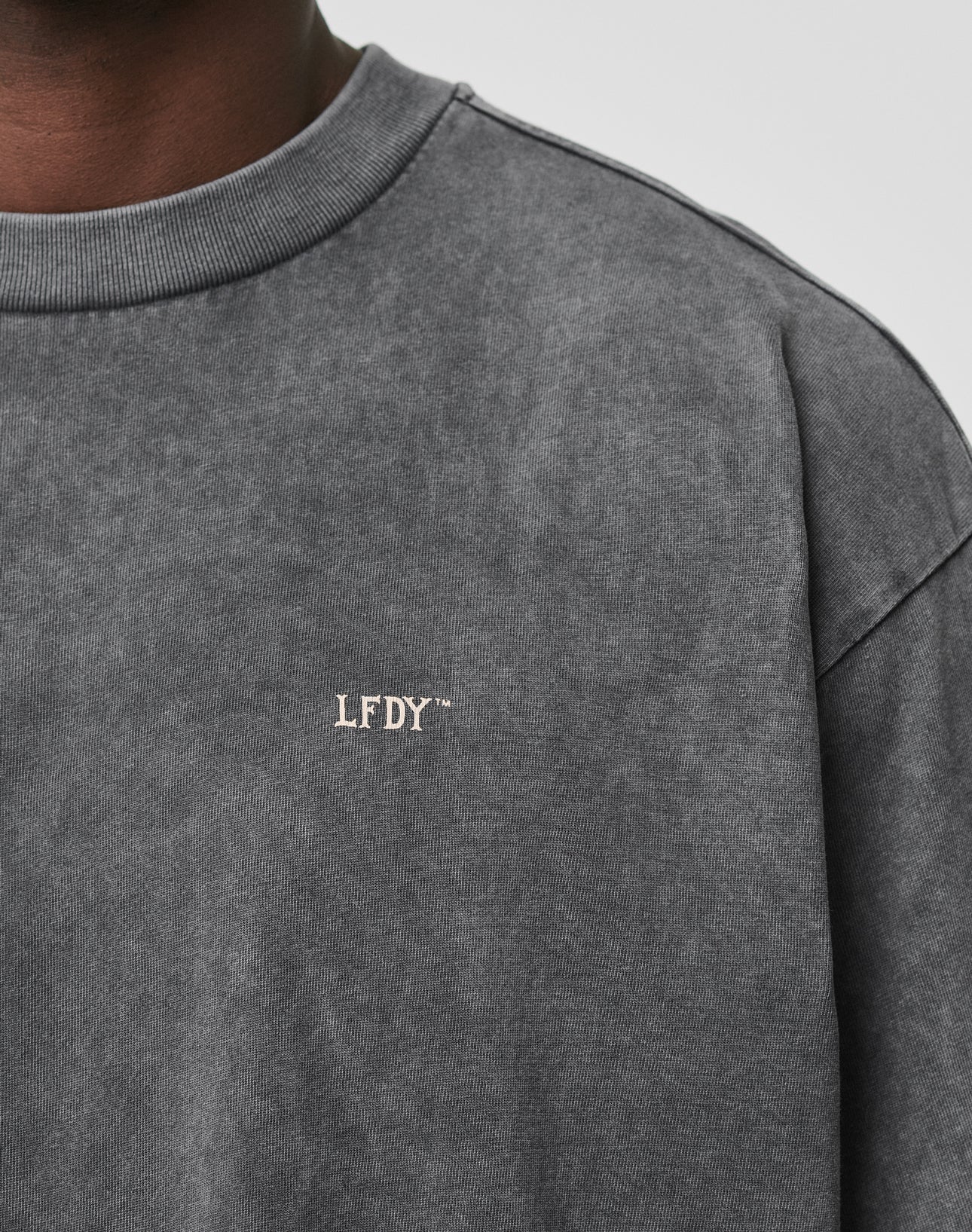 Close-up of a person wearing the LFDY Property Tee by LIVE FAST DIE YOUNG. The washed dark gray, oversized-fit shirt features small white LFDY™ embroidery on the left chest. Only the lower face and neck are partially visible.