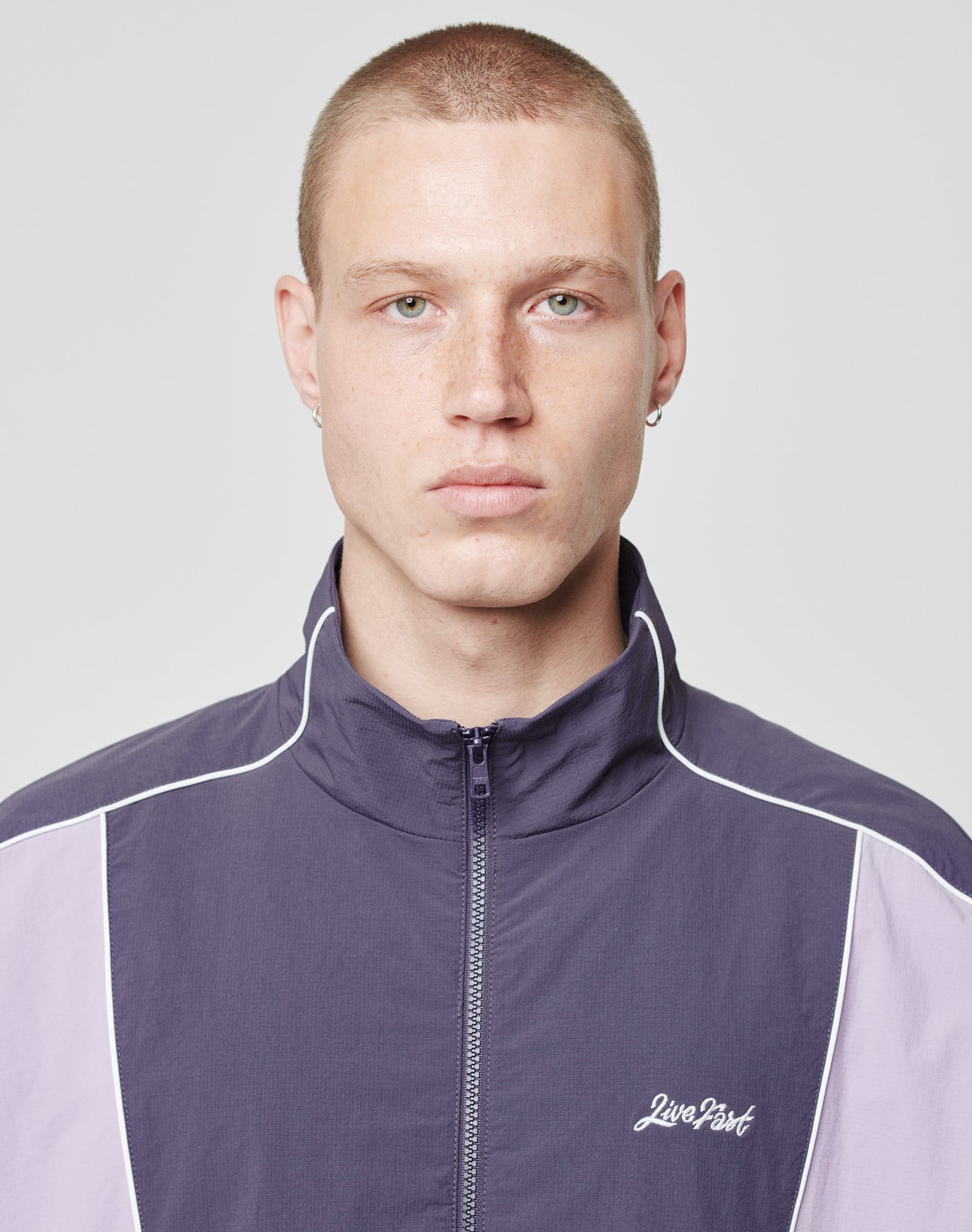 A person with short blond hair and light skin wears the LIVE FAST DIE YOUNG Basic Nylon Trackjacket in purple with white piping, logo patch, and side pockets, looking directly at the camera against a plain light gray background.