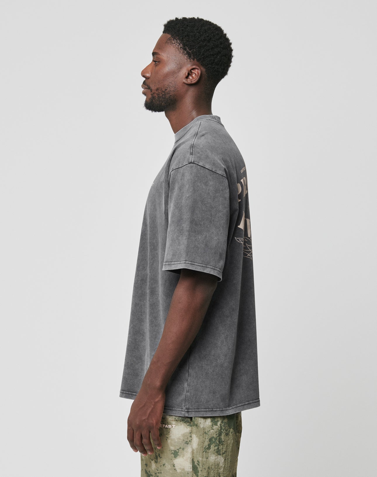A man with short curly hair and a beard stands in profile, wearing the LIVE FAST DIE YOUNG LFDY Property Tee—an oversized, faded gray t-shirt made from 100% Baumwolle—and green camo pants against a white background.
