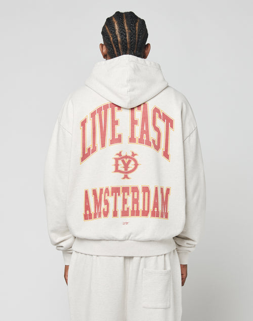 A person with braided hair is seen from behind wearing the LIVE FAST DIE YOUNG Live Fast Amsterdam Hooded in light gray, featuring an oversize fit and bold red and yellow LIVE FAST AMSTERDAM text with a small central logo front and back.